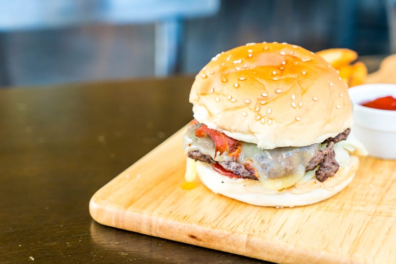 Gourmet cheeseburger with bacon and melted cheese on a wooden board, with fries and ketchup.