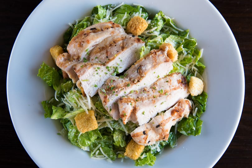 Overhead view of a Caesar salad with grilled chicken, croutons, and shredded cheese in a white bowl.