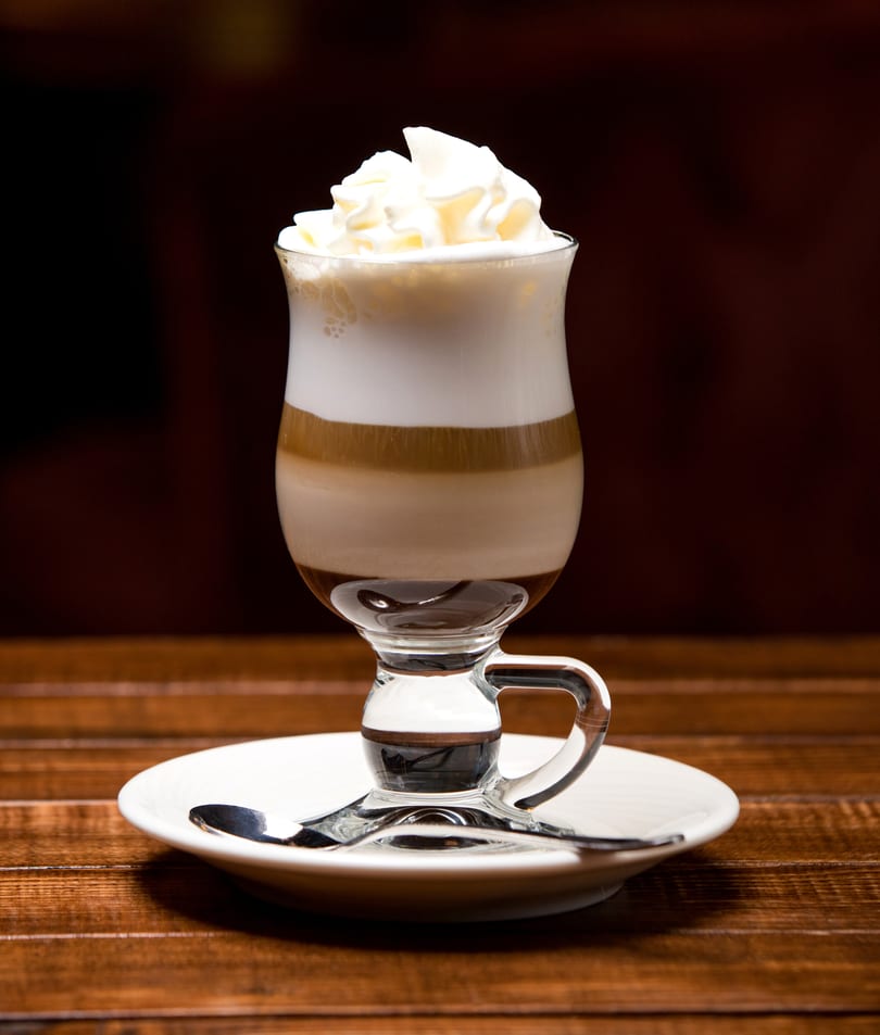 Layered coffee drink with whipped cream in a clear glass mug on a white saucer with a spoon.