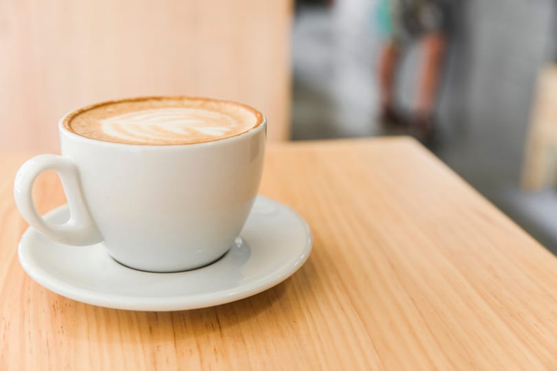 A white cup of latte with foam art sits on a saucer on a light wooden table.